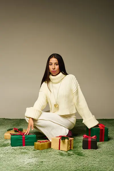 A stylish young woman sits on a green carpet, surrounded by colorful gifts and festive vibes. — Stock Photo