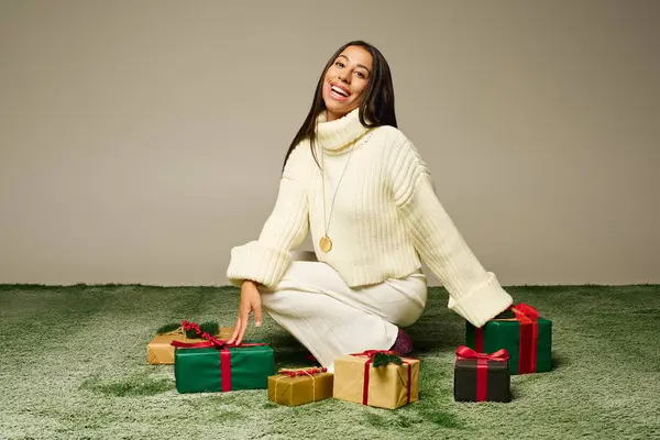 The woman sits on a soft surface, smiling brightly, with colorful presents around her. — Stock Photo
