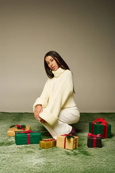 A stylish young woman kneels gracefully beside an assortment of colorful gifts, looking thoughtful. — Stock Photo