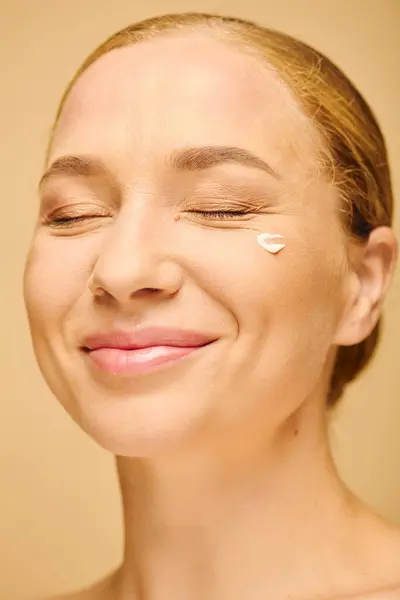 A youthful plus size woman smiles softly while posing with cream on her cheek in a warm space. — Stock Photo