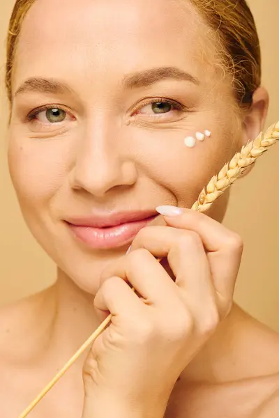 Una joven y hermosa mujer de talla grande se aplica crema en la mejilla mientras sonríe calurosamente. - foto de stock