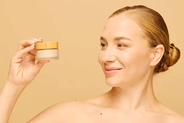 A radiant young woman smiles while showcasing a skincare product in a minimalist setting. — Stockfoto