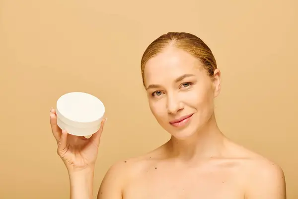 La mujer confiada sostiene un producto para el cuidado de la piel, sonriendo calurosamente sobre un fondo beige suave. - foto de stock