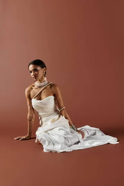 An elegant African American woman, dressed in a white gown and pearls, exudes grace against a brown backdrop. — Stock Photo