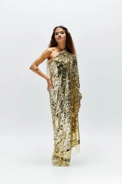 An elegant goddess in a shiny golden dress poses gracefully, radiating confidence in a serene studio space. — Stock Photo