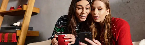 Zwei Freundinnen in festlichen Pullovern teilen Lachen und Freude, während sie Weihnachten feiern und aufs Smartphone schauen — Stockfoto