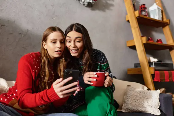 Two girlfriends celebrate the holidays, sharing laughter and warmth in cozy sweaters while looking at smartphone — Stock Photo