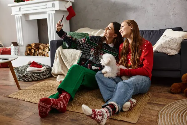 Two girlfriends in festive sweaters enjoy a joyful moment with their Bichon Frise while taking selfie — стоковое фото