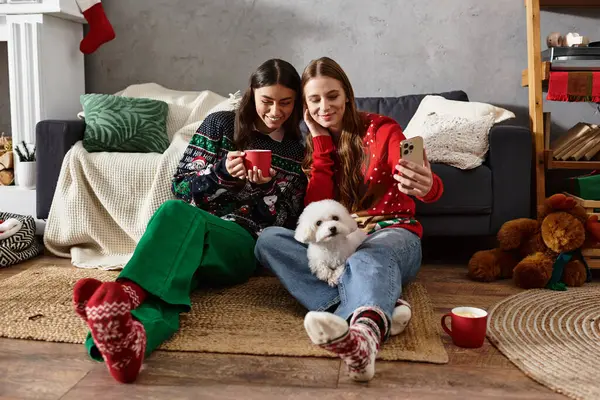 Girlfriends wearing cozy sweaters enjoy festive drinks and take selfie with their Bichon frise. — стоковое фото