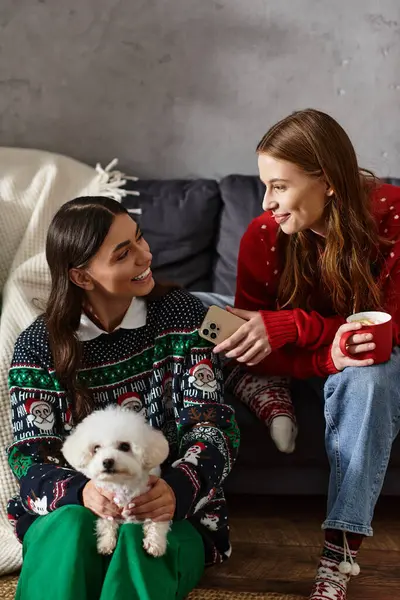 Two girlfriends joyfully celebrate the holiday season with a Bichon frise in their cozy living room. — Stock Photo
