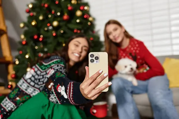 Two friends in festive sweaters enjoy a cozy Christmas gathering with their cute Bichon Frise dog. — Photo de stock