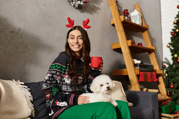 A young woman in festive attire enjoys the holidays with her adorable Bichon Frise by the cheerful Christmas decor. — Stock Photo