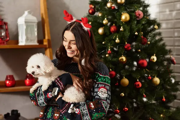 A young woman smiles brightly while celebrating New Year with her cute Bichon Frise beside the festive tree. — Stock Photo
