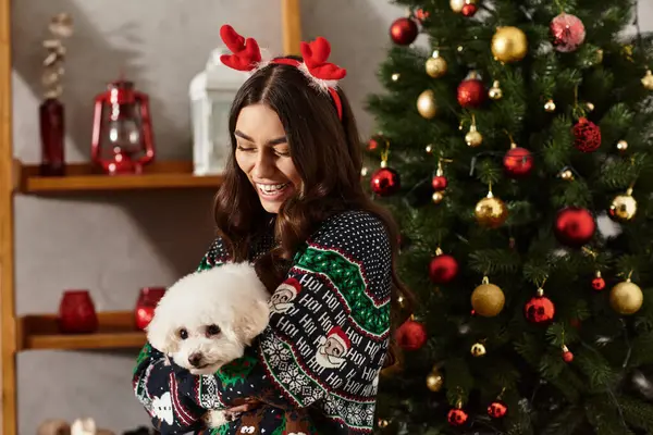 A young woman in a festive sweater smiles while celebrating the New Year with her adorable Bichon Frise dog. — Foto stock