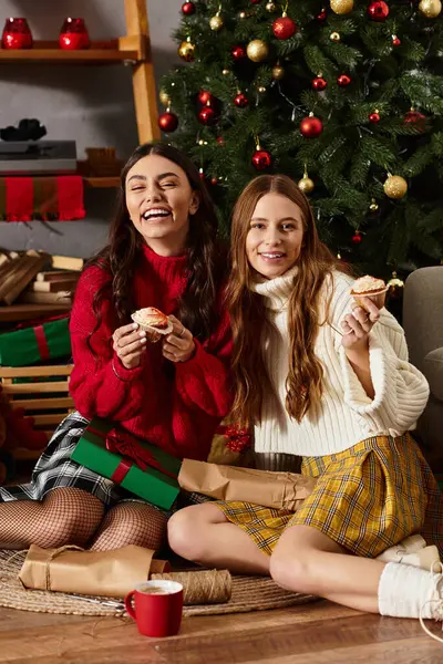 Girlfriends share laughter and festive treats together near a sparkling Christmas tree at home. — Stock Photo