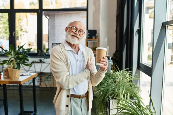 A senior businessman with a beard relaxes in a sleek office while enjoying a cup of coffee, radiating confidence. — Stock Photo