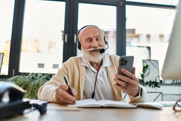 A senior businessman with a beard smiles while using his phone, effortlessly multitasking in a modern workspace. — Stock Photo