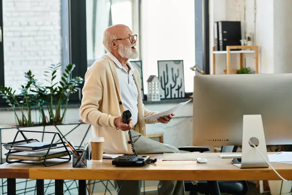 A senior businessman with a beard actively works in his modern office, balancing documents and a call. — Stock Photo