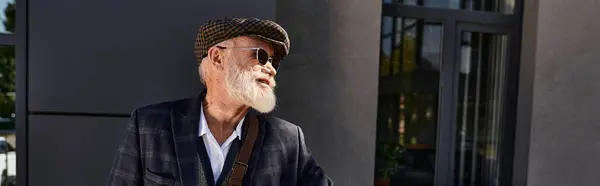 Un hombre mayor de moda luce una elegante chaqueta a cuadros y una gorra plana, disfrutando del día de otoño junto a un edificio moderno. - foto de stock