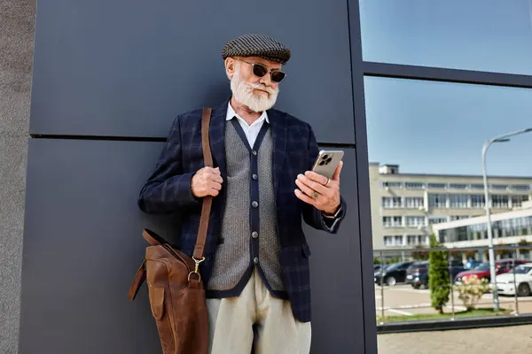 Un uomo anziano ben vestito si distingue casualmente da una struttura contemporanea, assorto nel suo smartphone durante l'autunno.. — Foto stock