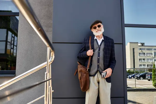 Un uomo anziano alla moda si trova con fiducia accanto a una struttura elegante, in mostra il suo vestito autunno con fascino. — Foto stock