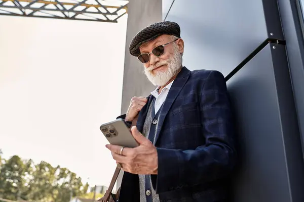 Ein modischer älterer Mann mit Bart steht lässig in der Nähe einer modernen Struktur und checkt sein Telefon. — Stockfoto