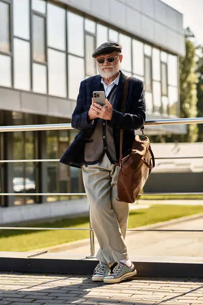 Un uomo anziano alla moda con la barba si trova accanto a una struttura elegante e contemporanea, controllando il suo smartphone.. — Foto stock