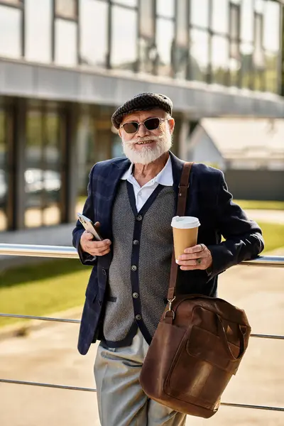 A stylish senior man with a beard stands confidently by a modern building, sipping coffee as autumn arrives. — Stock Photo