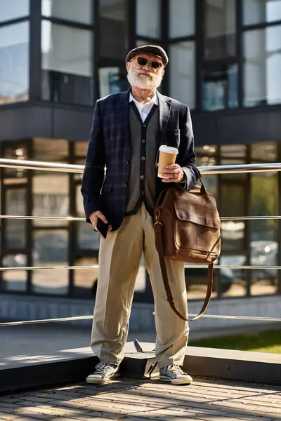 Un uomo anziano alla moda con la barba tiene un caffè e si distingue con fiducia da una struttura contemporanea in autunno.. — Foto stock