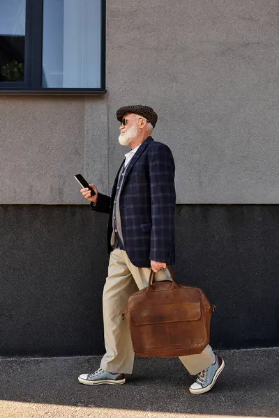 A fashionable senior man walks with a bag, dressed in smart casual attire, near a contemporary building in autumn. — Stock Photo