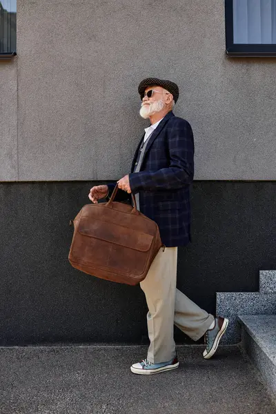 A stylish senior man in a checkered blazer and flat cap walks with his bag by a modern building in autumn. — Stock Photo
