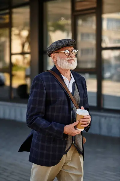 A dapper senior man in a checkered blazer strolls with coffee, embracing autumns charm by modern buildings — Stock Photo