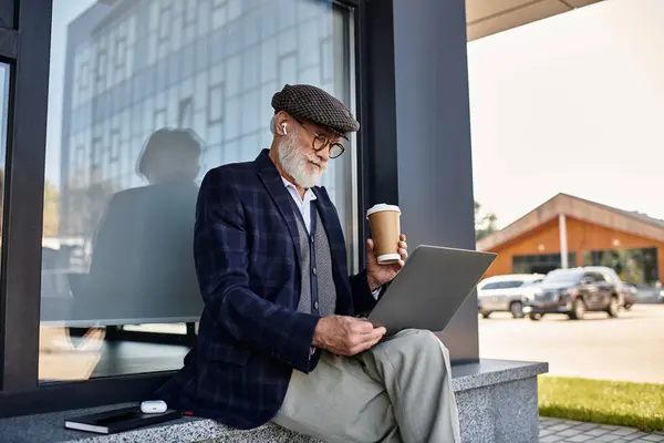 Un uomo anziano alla moda si siede vicino a una struttura contemporanea, sorseggiando caffè e concentrandosi sul suo computer portatile in autunno.. — Foto stock