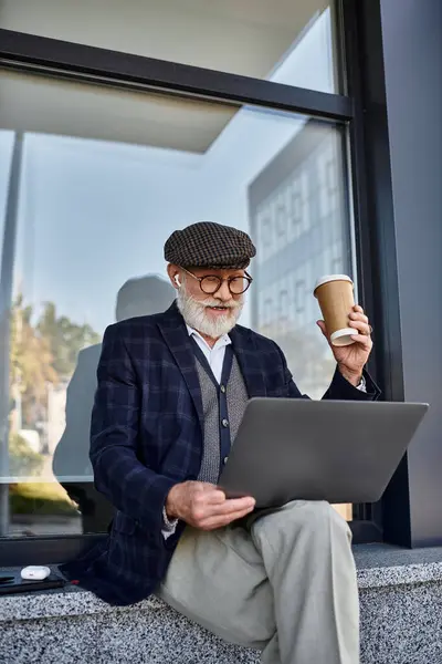 Un homme âgé élégant avec une barbe sirote du café et travaille sur son ordinateur portable par un bâtiment moderne. — Photo de stock