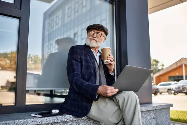 A fashionable senior man sips coffee, working on his laptop, dressed in a checkered blazer and flat cap. — Stock Photo