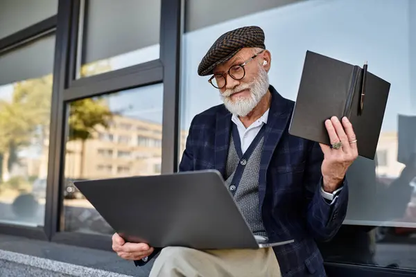 Un uomo anziano alla moda studia il suo computer portatile mentre seduto fuori da una struttura contemporanea in autunno. — Foto stock