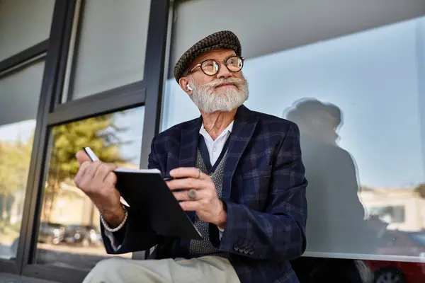 A fashionable senior man sits thoughtfully outside a modern building, enjoying the autumn breeze as he sketches. — Stock Photo