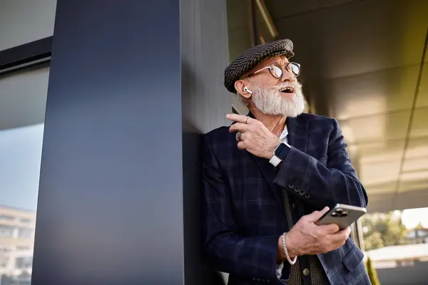 A stylish senior man in a checkered blazer and flat cap leans against a sleek building, embracing autumn. — Stock Photo