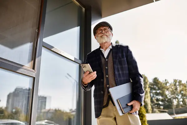 A fashionable senior man stands confidently by a modern building, holding a phone and documents during autumn. — Stock Photo