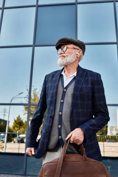 A fashionable senior man stands confidently near a contemporary building, sporting a beard and cap in autumn. — Stock Photo