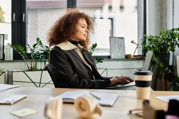 A woman with curly hair intently works on her laptop in a vibrant office filled with greenery. — Stock Photo