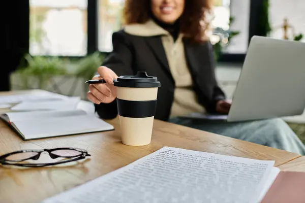 A woman with curly hair sits at a wooden desk, smiling as she reaches for her coffee cup. — Stock Photo