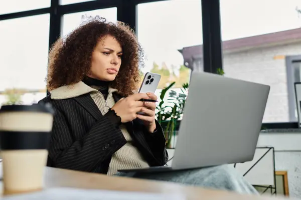 Une femme aux cheveux bouclés interagit avec son smartphone tout en travaillant sur un ordinateur portable. — Photo de stock