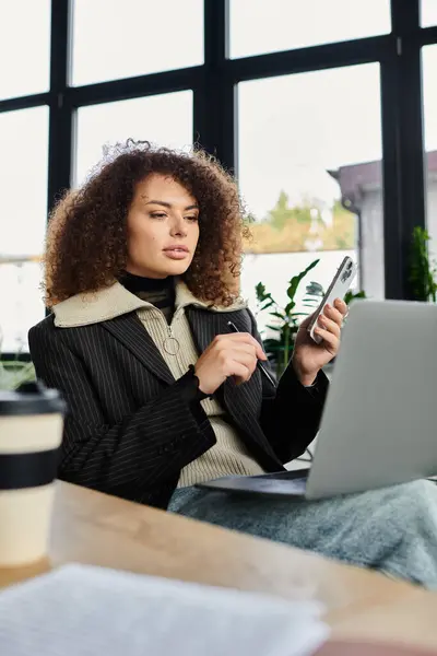 A woman focused on her tasks uses a laptop and phone in a sunlit workspace. — Stock Photo