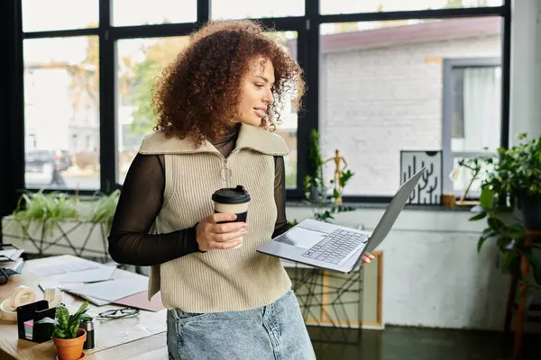 Une femme s'engage avec son ordinateur portable, tenant une tasse de café dans un bureau contemporain rempli de verdure. — Photo de stock