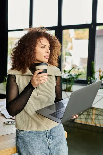 A woman holds a coffee cup and focuses on her laptop in a lively setting filled with plants. — Stock Photo