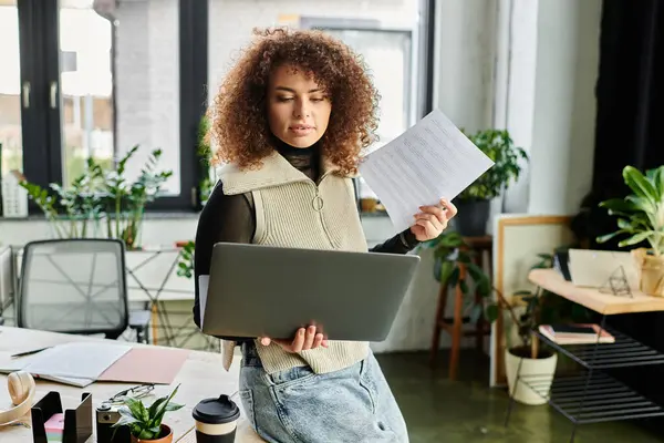 A woman holds documents while working on a laptop surrounded by greenery in a bright workspace. — Stock Photo