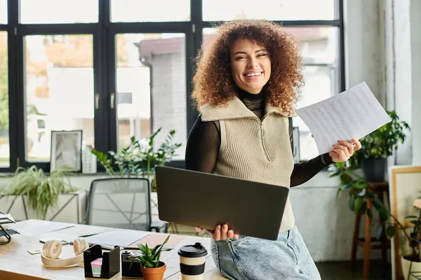A happy woman stands in a bright office area, holding a laptop and paperwork while smiling. — Stock Photo