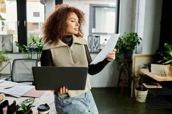 A woman in a cozy workspace studies important papers while holding a laptop and looking engaged. — Stock Photo