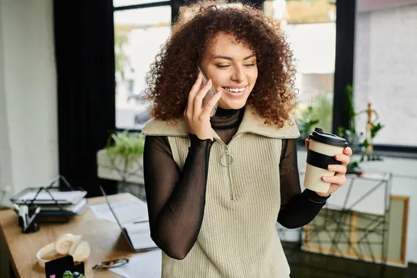 A cheerful woman talks on her phone while sipping coffee in a cozy, well lit office. — Stock Photo